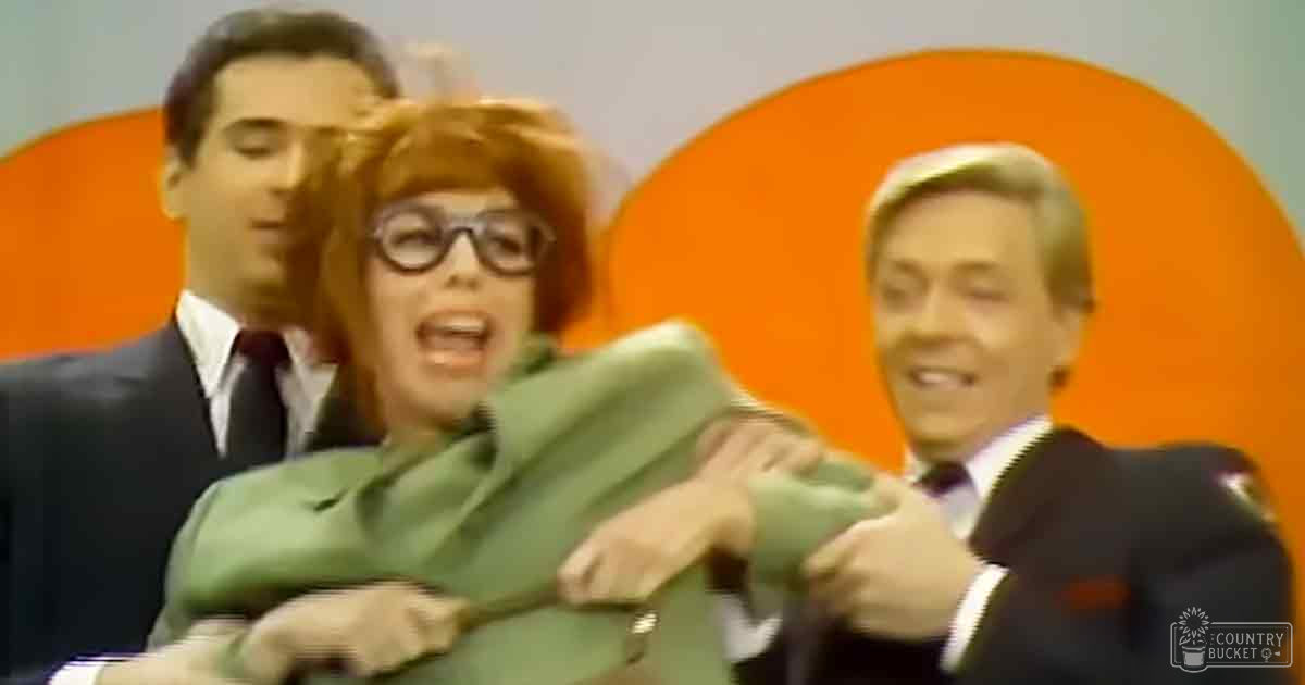 A woman in a green suit stands at center, flanked by two men in formal attire; an orange circular backdrop suggests a stage setting.