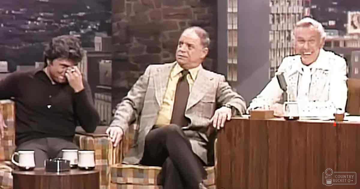Three individuals seated at a talk show set against an urban cityscape backdrop, with patterned chairs and wooden desk, suggesting a nighttime studio environment.