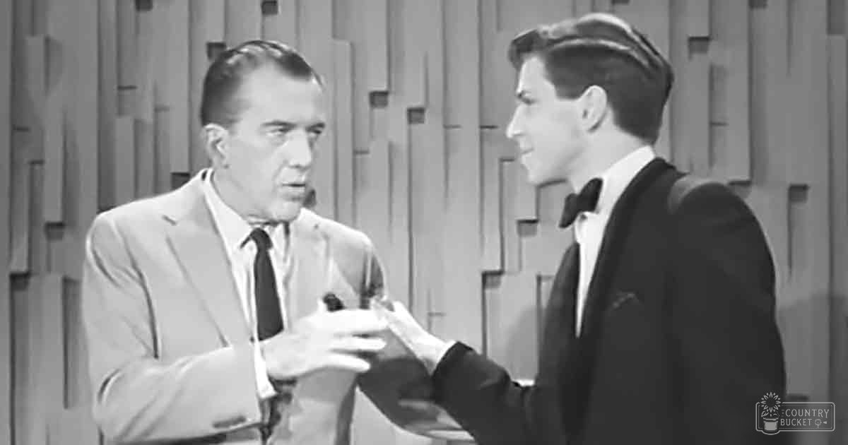 A mid-20th century studio scene with two middle-aged men dressed formally, one holding a microphone against a geometrically patterned wall backdrop.