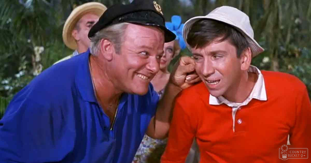 Tropical outdoor setting with palm trees and lush greenery, with a middle-aged male wearing a blue polo shirt and a captain's hat, interacting with a young adult male in an orange polo shirt and white bucket hat.