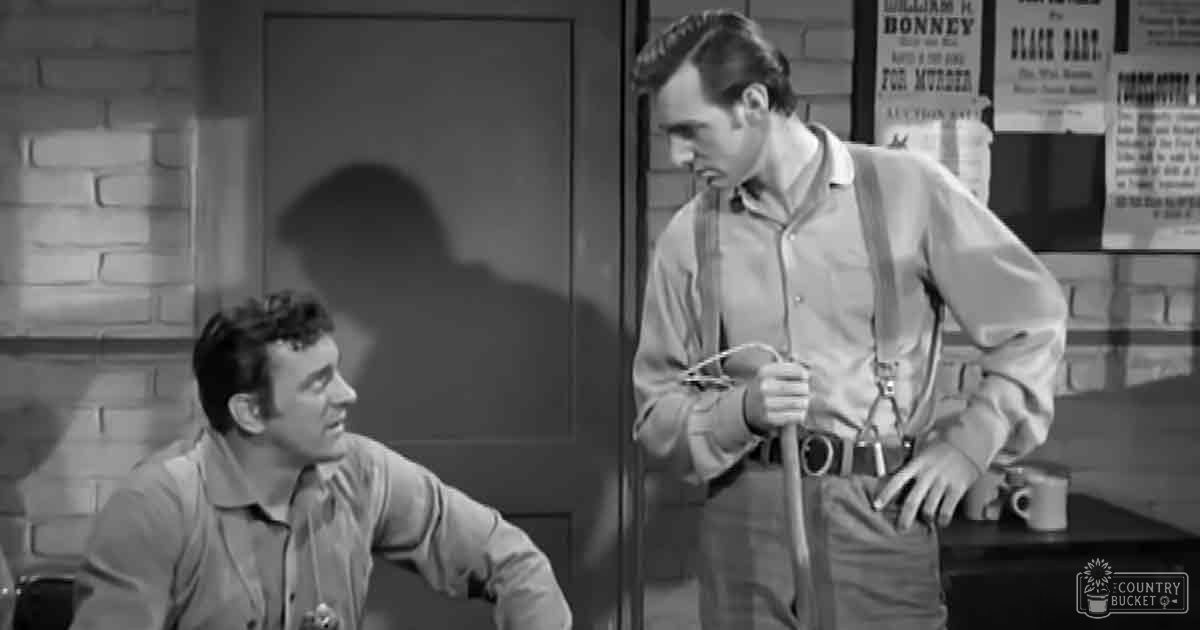An old Western film scene set in a saloon or sheriff's office with two adult males wearing button-up shirts and ties, surrounded by wooden furniture and posters referencing characters like William H. Bonney and Black Bart.