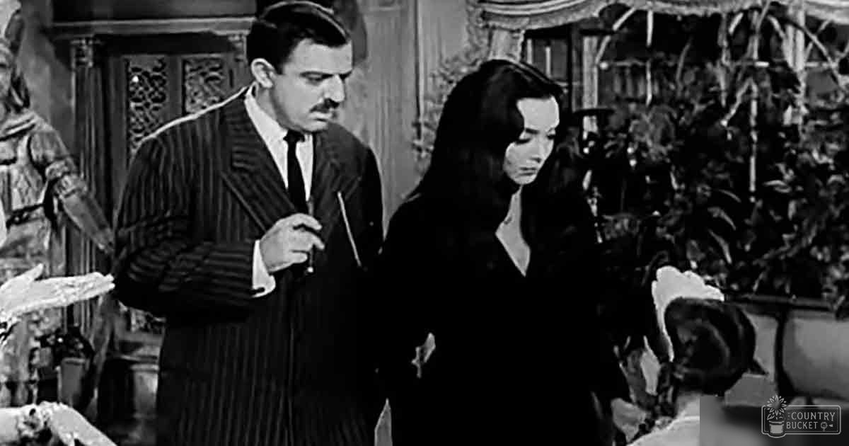 An adult male wearing a striped suit jacket with a white shirt and dark tie, seated indoors alongside an adult female in a dark-colored dress with long sleeves; ornate furniture suggests a formal room setting, likely daytime with natural light.