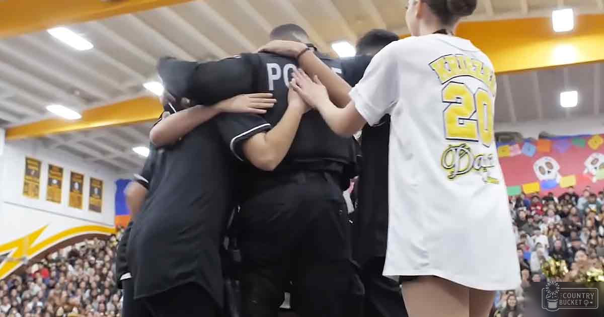 A group huddle in an indoor sports arena with young adults and teens wearing dark athletic uniforms, surrounded by a crowd of spectators under fluorescent lighting.