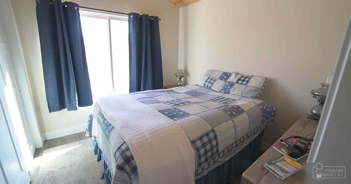 A bedroom with a patchwork quilt in shades of blue and white, dark curtains allowing natural light to enter, and furniture including a nightstand with a lamp and books or magazines.
