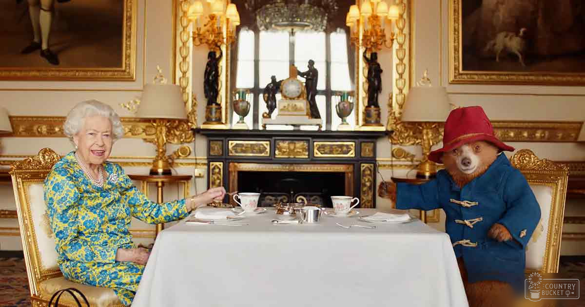 A woman with a brown bear, likely Paddington Bear, seated at an ornate table in a grand, formal setting with opulent gold and black furniture and bright natural light.