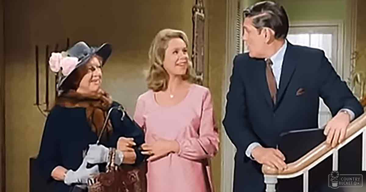 Indoor scene from a mid-20th century television show, with adult females and males dressed in period attire with pink dresses, dark coats, and a staircase leading upwards.