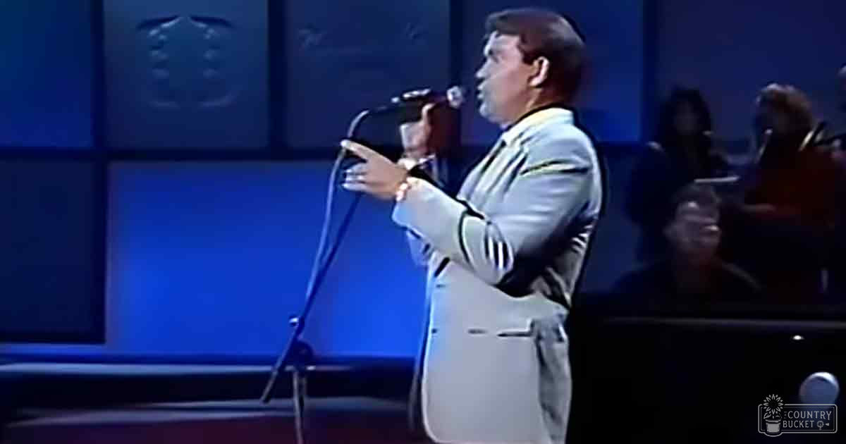 An adult male singer in a light-colored suit with vertical stripes, singing into a microphone on a blue-lit stage with soundproofing panels and part of an audience seated in the background.