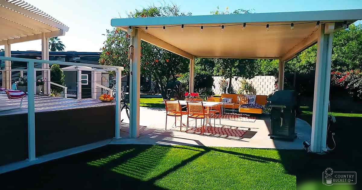 A residential backyard patio area with outdoor furniture, pergola structure, string lights, artificial grass, bowl of fruit, and a small shed or storage unit surrounded by trees and shrubs. The setting appears to be during spring or summer with clear skies.