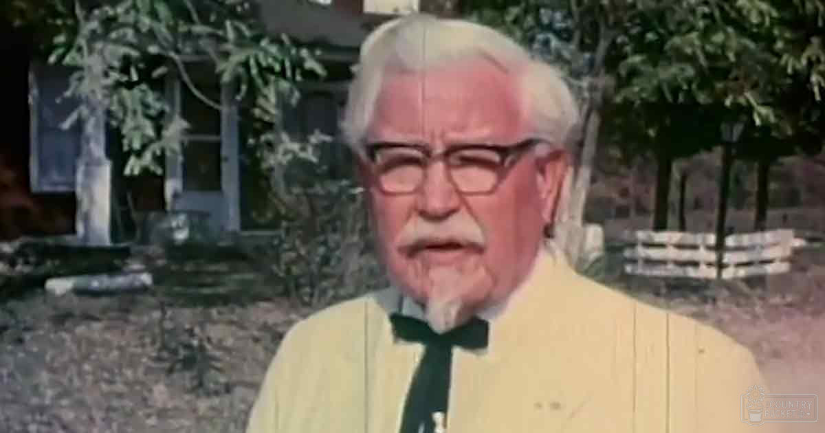 An older adult male in a rural setting, wearing a tailored light yellow suit jacket with a black bow tie and standing amidst greenery and a rural house.