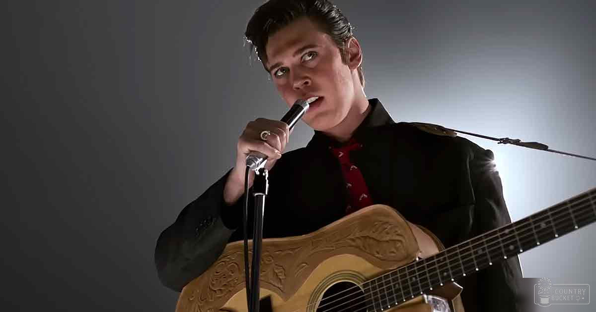 A young adult male wearing a dark blazer with red shirt and patterned tie, performing on stage in an indoor music venue under professional lighting.