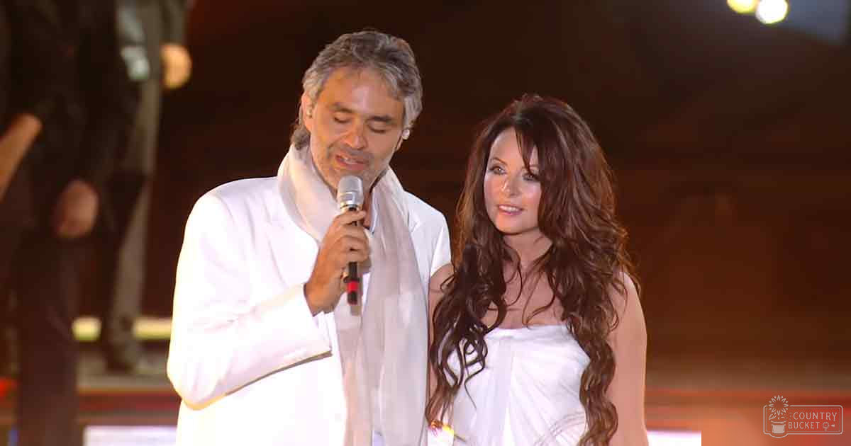 A middle-aged male and an adult female performing on stage with white attire against dim lighting; microphone visible, suggesting a concert or theatrical setting.