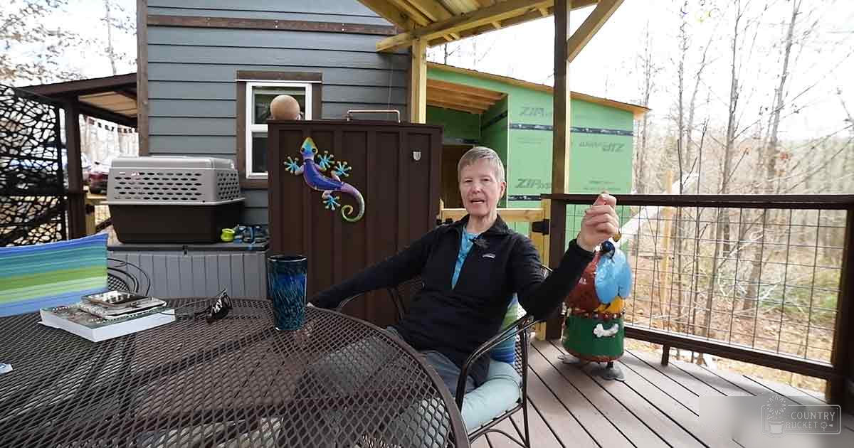 An older adult male sits at a metal patio table with a woven chair, surrounded by greenery and decor in what appears to be an early spring or late fall backyard setting.