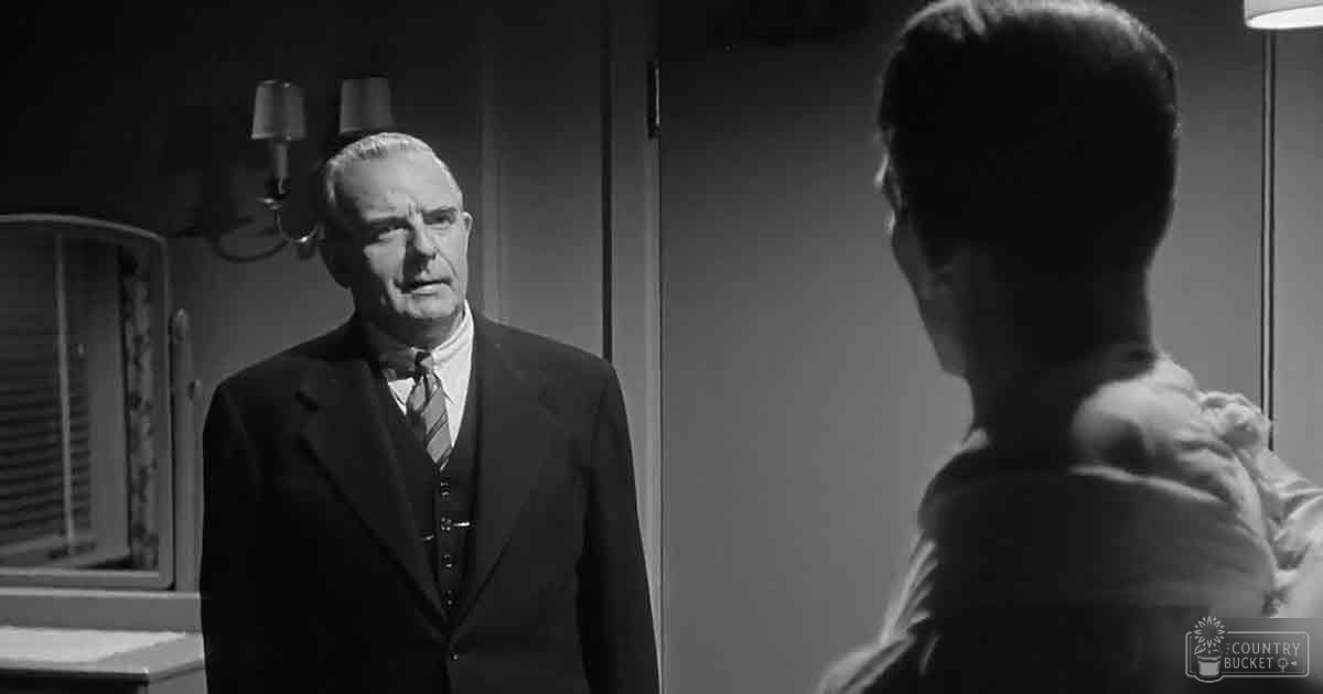 An older adult male wearing a dark suit jacket, light-colored shirt, and striped tie stands in an indoor professional setting with wall-mounted lamps above his head. The scene suggests mid-20th century decor.