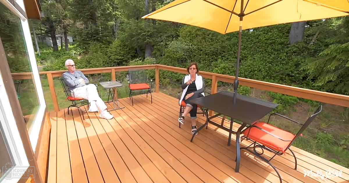 Two older adults sitting on a wooden deck in an outdoor patio area surrounded by lush greenery, wearing casual attire and sunglasses under the shade of a yellow umbrella.