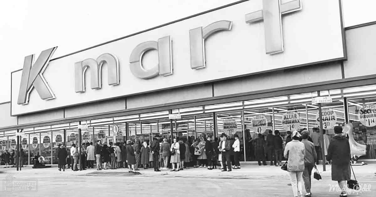 An adult male singer wearing a dark jacket and light-colored pants stands outside a Kmart store, while an older female shopper in a fur-lined coat walks by. A large 'Kmart' sign is prominently displayed above the entrance.