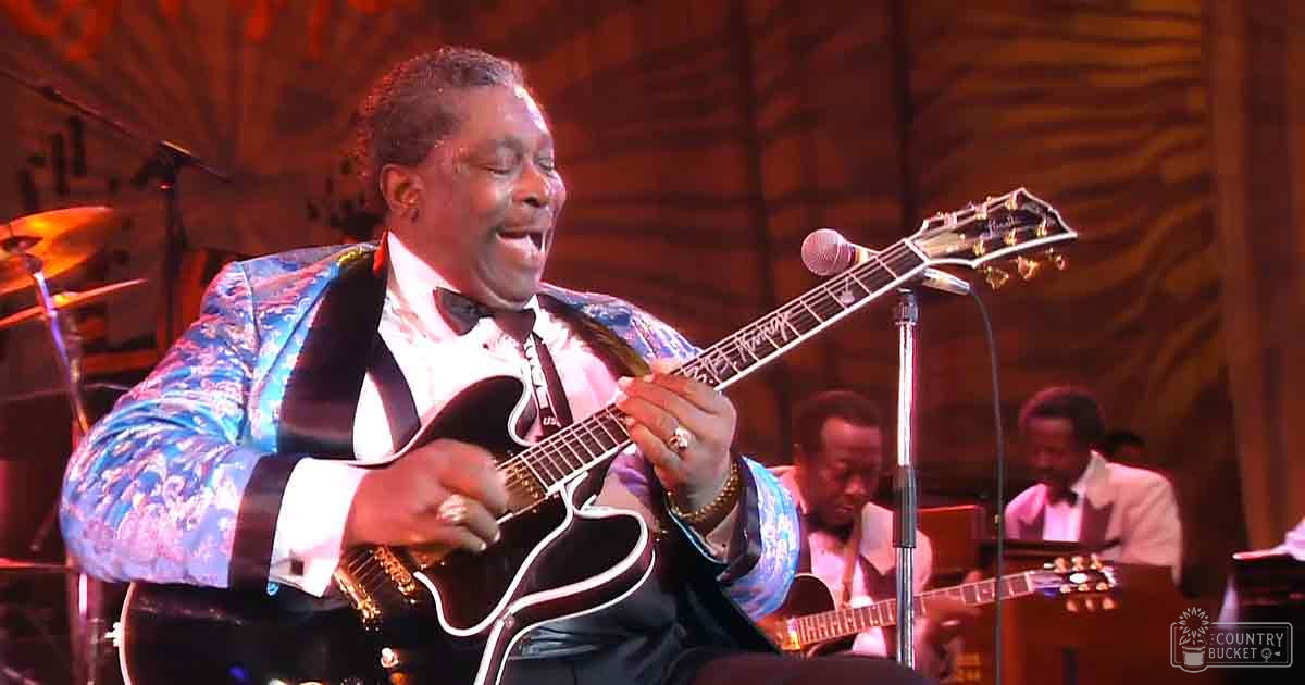 A male musician wearing a patterned blue and white jacket performs on stage at an indoor concert venue, surrounded by other musicians with microphones and musical equipment.