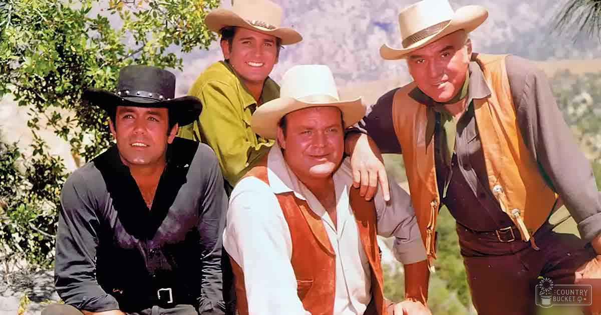 A group of actors from 'The Dukes of Hazzard' dressed in cowboy attire, standing outdoors with trees and hills in the background on a sunny day.