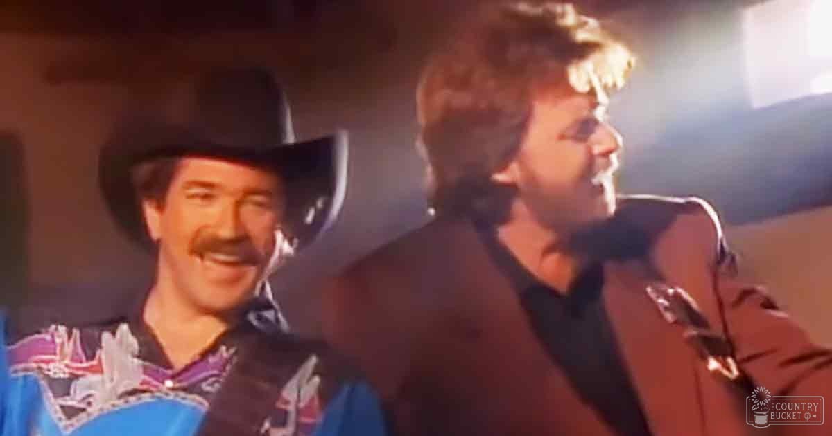 Middle-aged male singer in a cowboy shirt with intricate embroidery and blue bandana performing alongside an adult female in a dark jacket, possibly leather, on stage under warm lighting.