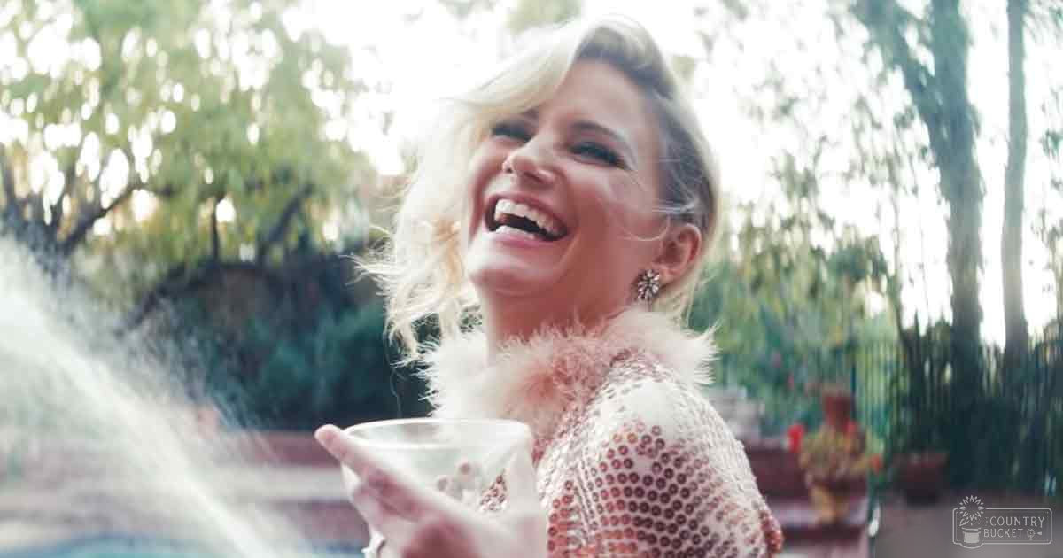 A young adult female in a textured white garment with a rounded neckline, possibly inspired by the 1920s-1930s era, holding a white bowl outdoors in what appears to be a garden or park with trees and greenery.