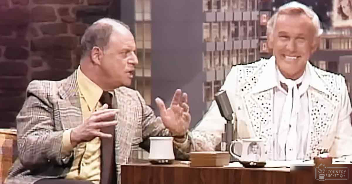Indoor studio talk show setting, middle-aged man plaid blazer yellow shirt, older adult white vest black polka dots, wooden table mugs papers books microphone shelves decorative items stone wall casual professional atmosphere bright even lighting earthy tones brown white yellow black accents