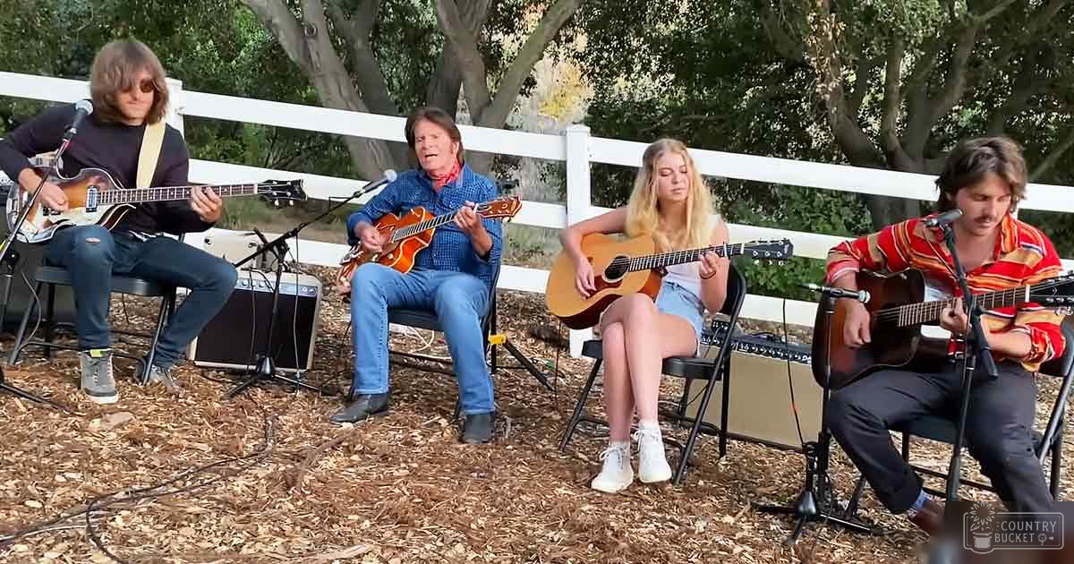 Four musicians performing outdoors on black chairs with microphones and amplifiers; trees and a white picket fence indicate an outdoor garden or park setting under natural daylight.