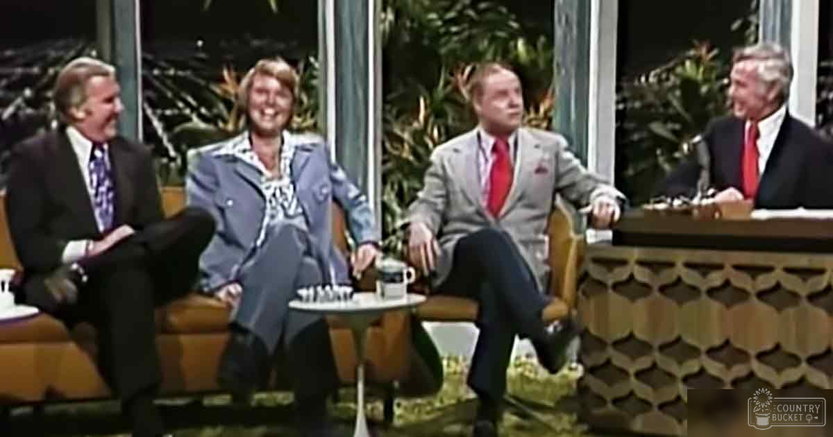 A talk show set with four individuals seated around a table indoors, with large windows with greenery visible outside and furniture from an earlier era.