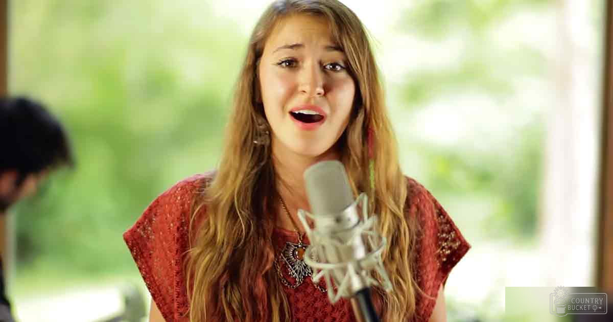A young adult female singer wearing a red top with large hoop earrings, performing into a microphone in an indoor setting with natural light streaming through windows.
