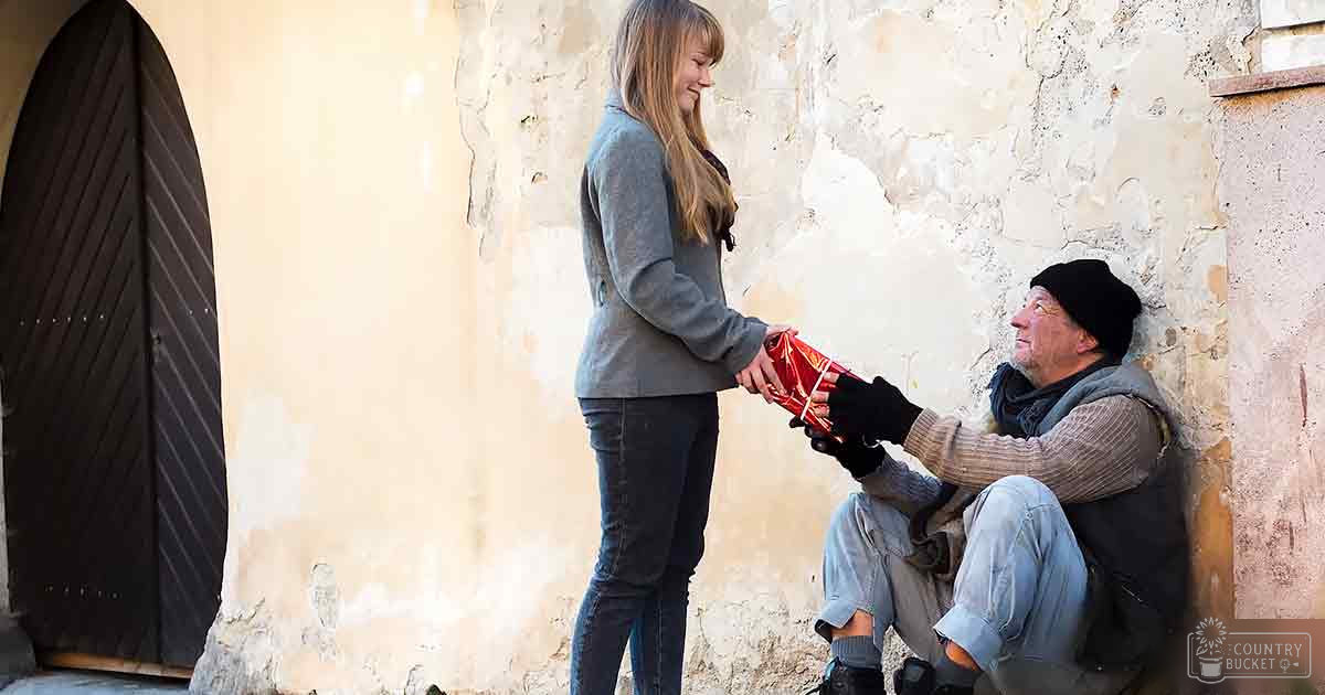 A young adult female in a gray long-sleeve sweater hands a wrapped gift to an older adult male wearing a black beanie and light brown knitted sweater, set against a weathered urban wall.