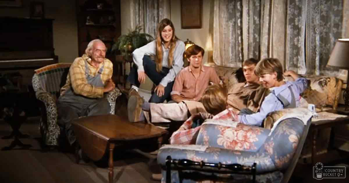 A family gathering in a vintage-style living room with floral-patterned armchair, wooden coffee table, piano, and curtains.