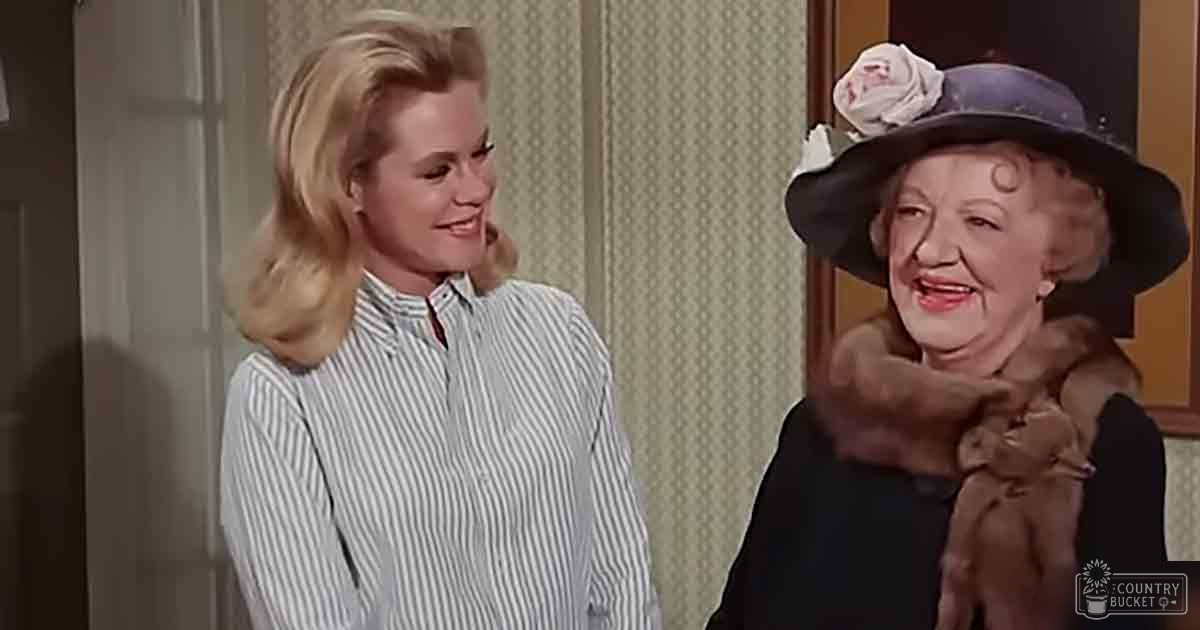 A young adult female wearing a white and gray striped shirt stands next to an older adult female dressed in a dark-colored dress with fur trim, both within a mid-20th-century styled interior setting.