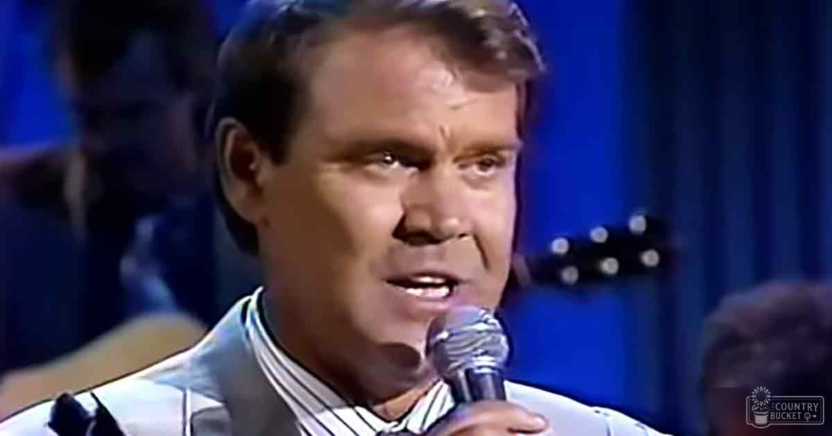 A middle-aged male singer wearing a light-colored tailored suit with striped shirt and dark tie, performing into a microphone in a studio setting under blue lighting.