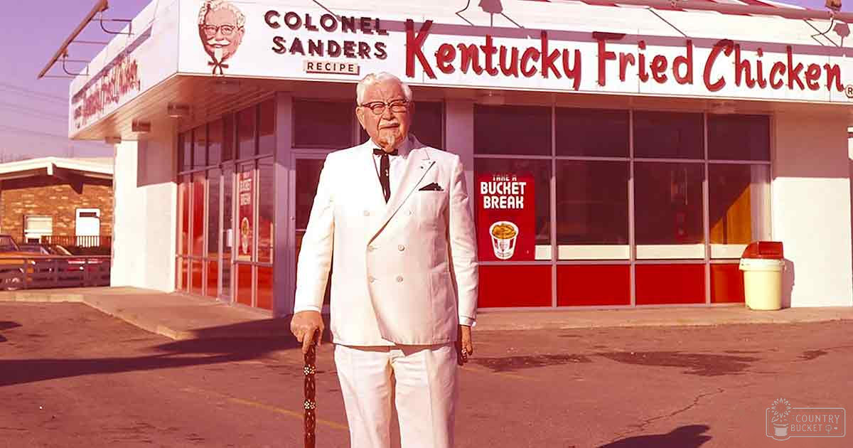 Colonel Sanders in a white suit with black tie and cane stands outside Kentucky Fried Chicken restaurant signage visible.