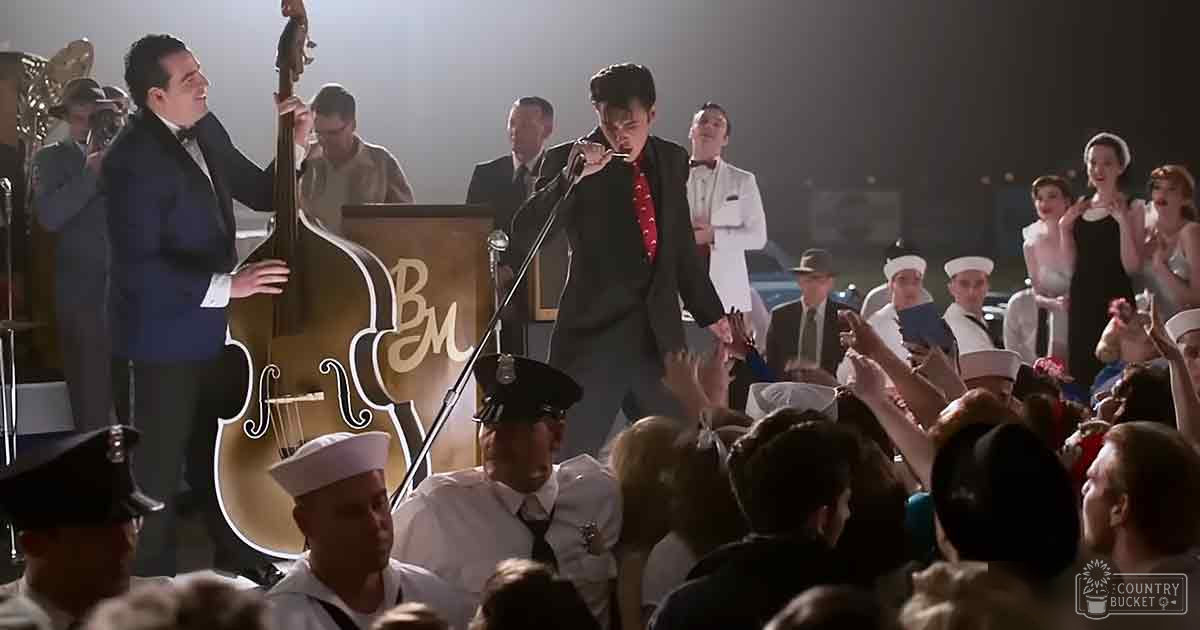 An adult male singer in a black tuxedo with a red cravat plays into a microphone on stage, surrounded by musicians and an audience in mid-20th century attire.