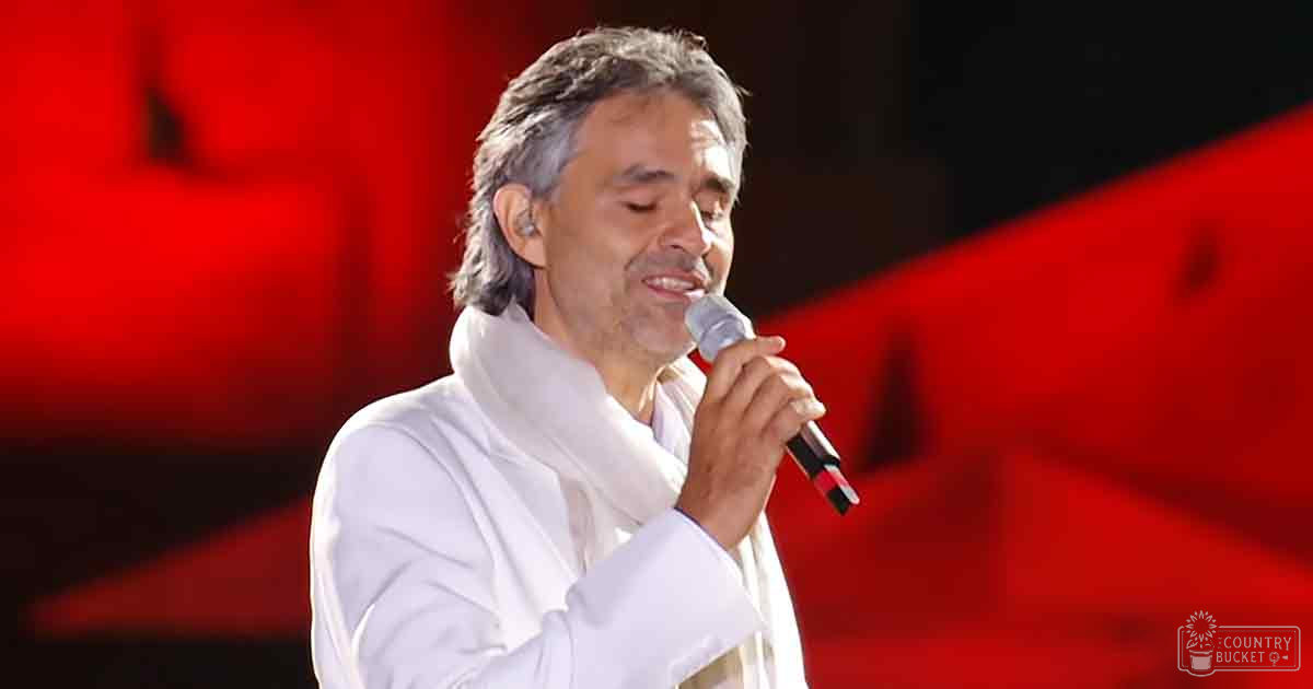 A middle-aged adult male singer wearing a loose, long-sleeved white garment with no visible pattern stands on stage under red lighting in what appears to be an indoor venue.