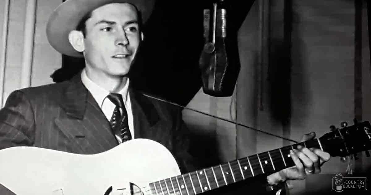 Young adult male in a fitted suit jacket with tie, holding an acoustic guitar in what appears to be a recording studio setting.