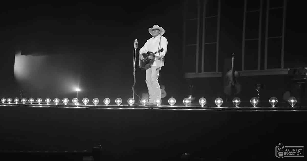 An adult male singer wearing a white button-down shirt and cowboy hat performs on stage in an indoor concert venue with dark curtains and seating, likely evening lighting.