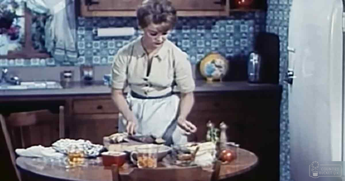 A young adult female in a kitchen setting, wearing a white apron over a light-colored dress with simple cut and straight neckline.