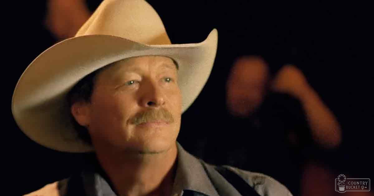 An older adult male in a Western setting, wearing a wide-brimmed cowboy hat and light-colored shirt at night.