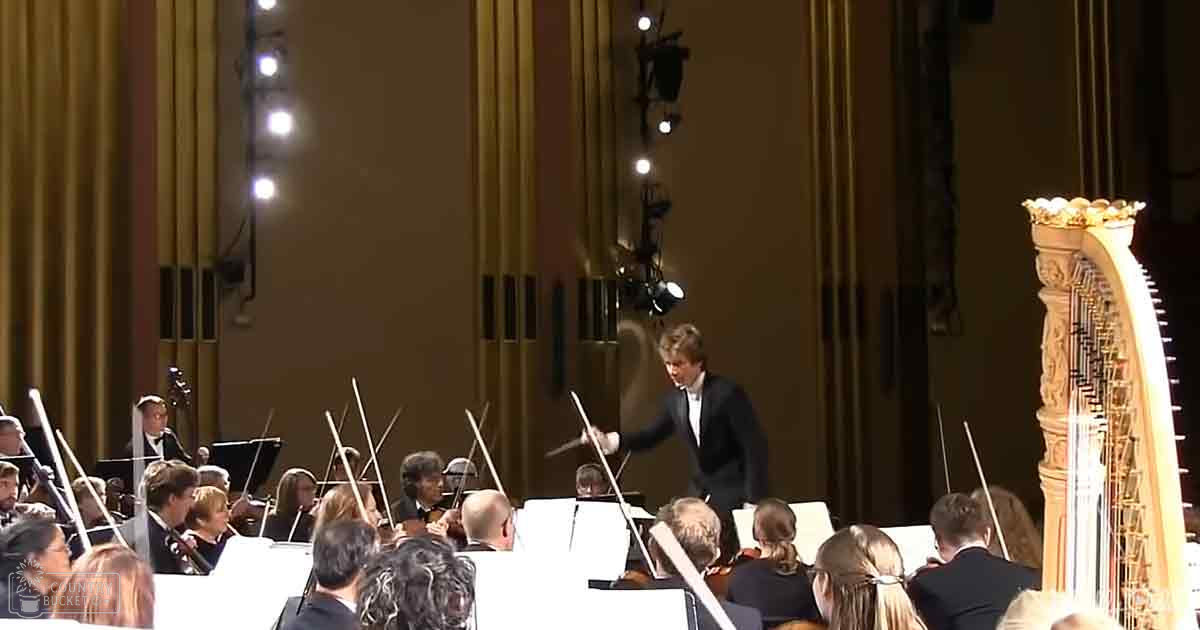 An adult male conductor leading an orchestra in a formal concert hall, with musicians dressed in black suits playing string instruments under artificial stage lighting.