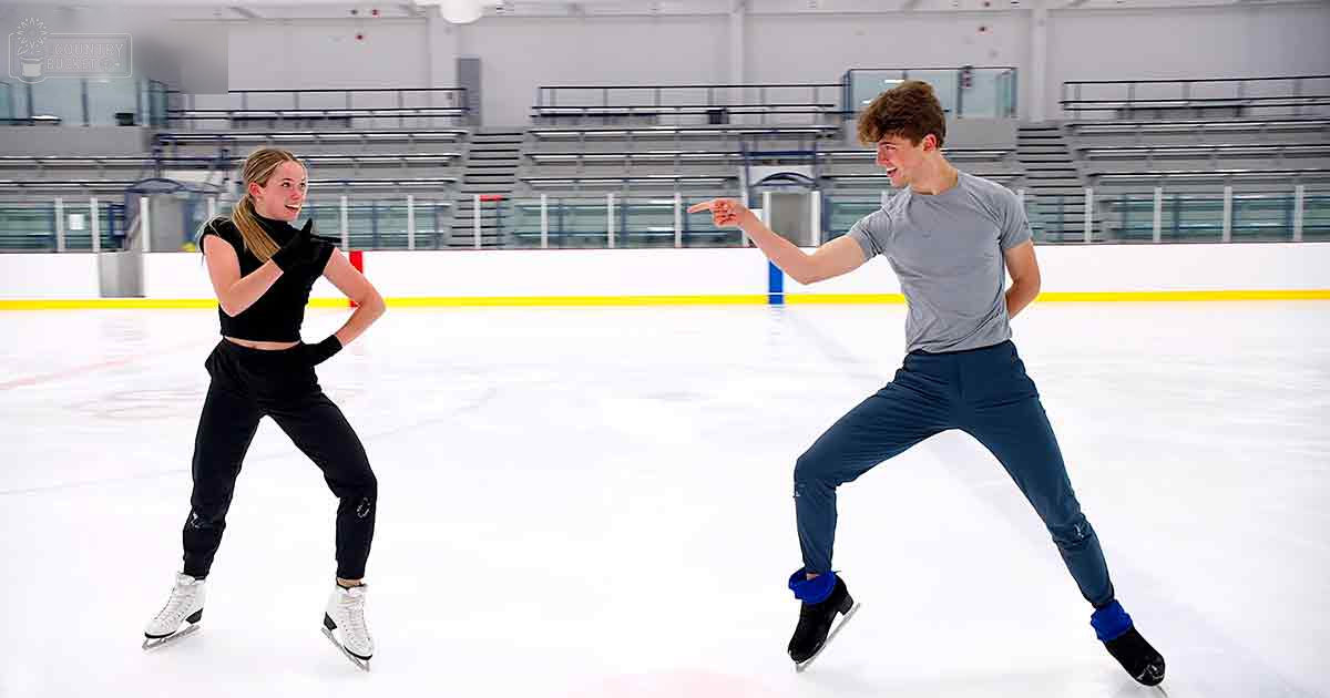 Young adult couple skating indoors at an ice rink with yellow boundary lines, surrounded by bleachers and natural light.
