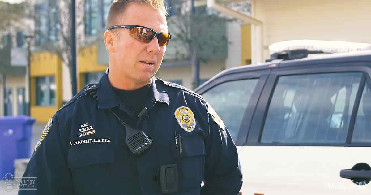 An adult male police officer in a dark blue uniform with 'M. BROUILLETTE' and 'SWAT' insignia, wearing sunglasses, standing on an urban street with greenery and trees in the background during daytime.