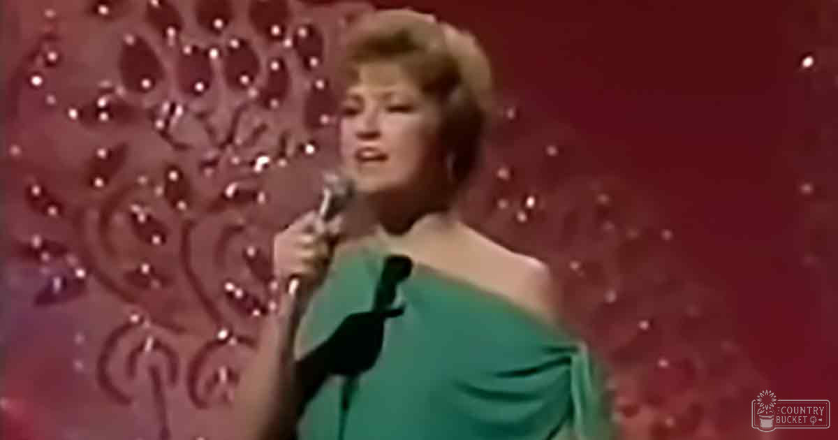 An adult female performer in a teal off-the-shoulder dress with draped design and fitted bodice, holding a microphone against a red patterned backdrop at an indoor performance venue.