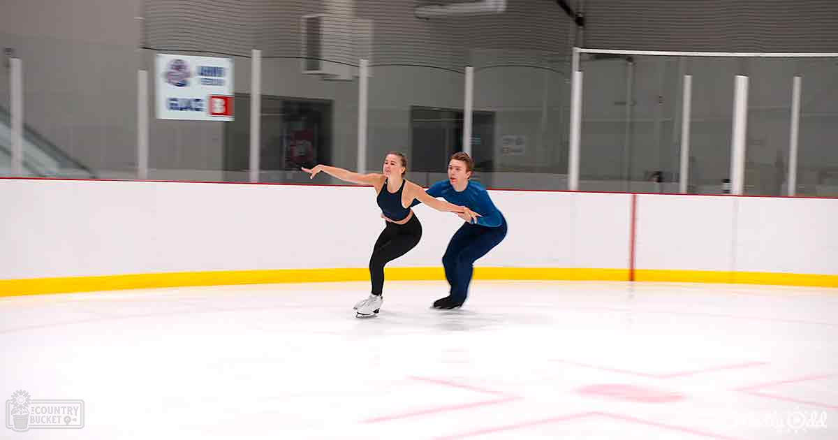 A figure skating pair practicing in an indoor ice rink with white barriers, yellow safety lines, and well-lit surroundings.