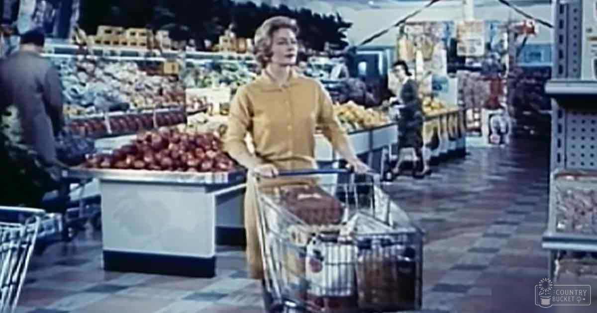 An adult female in a yellow button-up dress with long sleeves, walking inside a mid-20th century-style supermarket filled with grocery items like apples on display and shelves of bread.