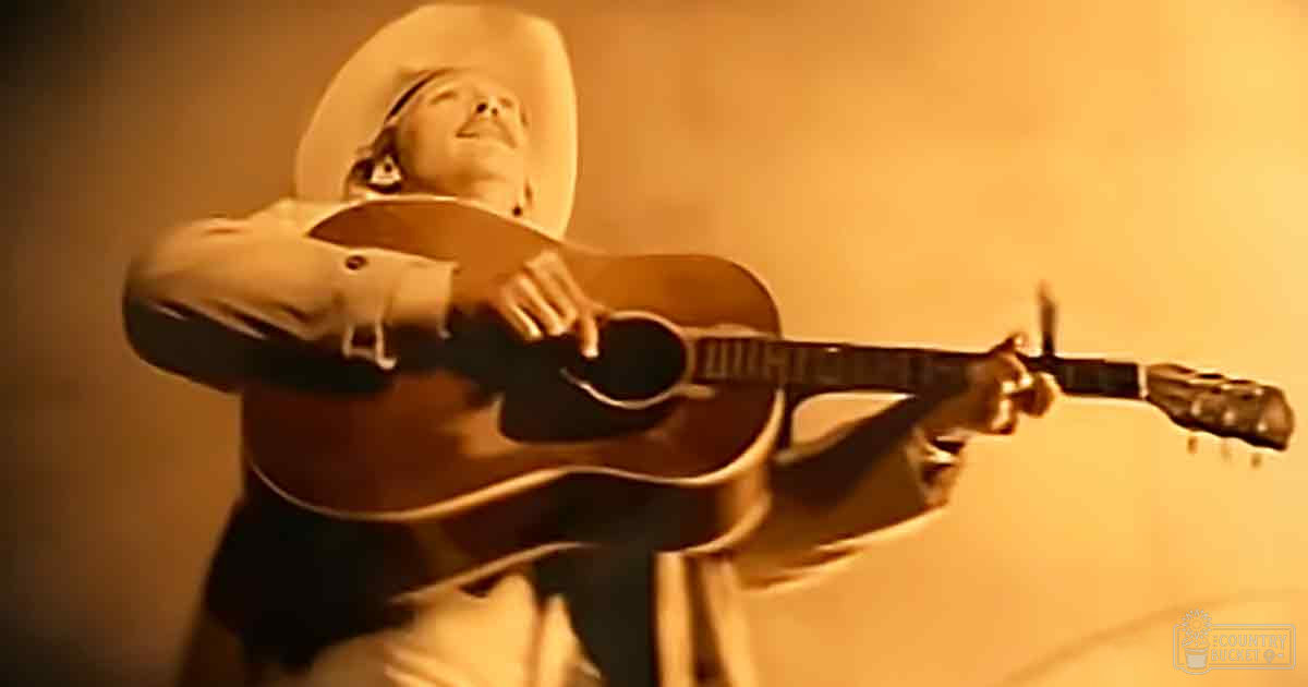 Adult male musician in a warm, indoor setting with sepia tones holding an acoustic guitar.
