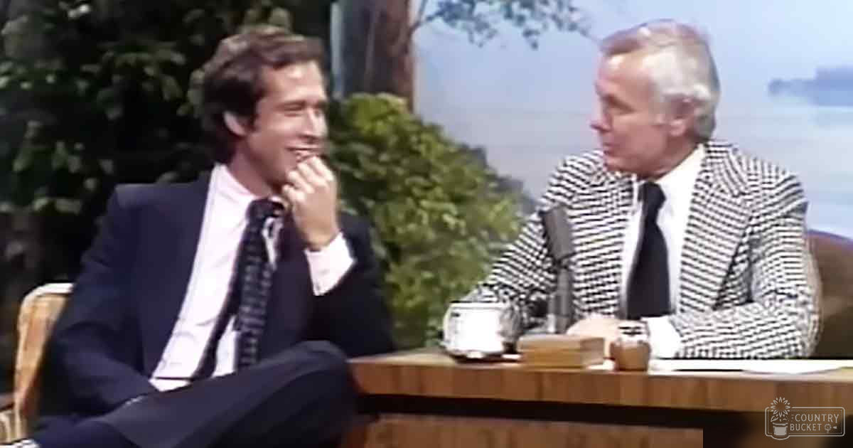 Adult male talk show host interviewing an older adult male guest in an outdoor studio setting with greenery and daytime lighting.