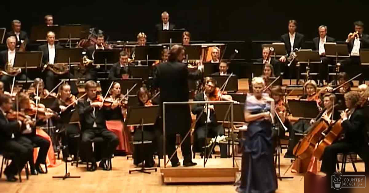 Adult male conductor leading an orchestra in a formal concert hall setting, surrounded by musicians dressed in dark formal attire under evening lighting.