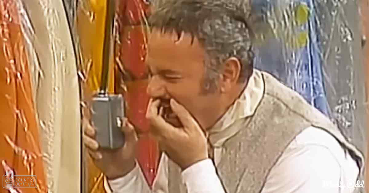 A middle-aged adult male wearing a white shirt with a light-colored vest over it, holding an old-fashioned tape recorder indoors.