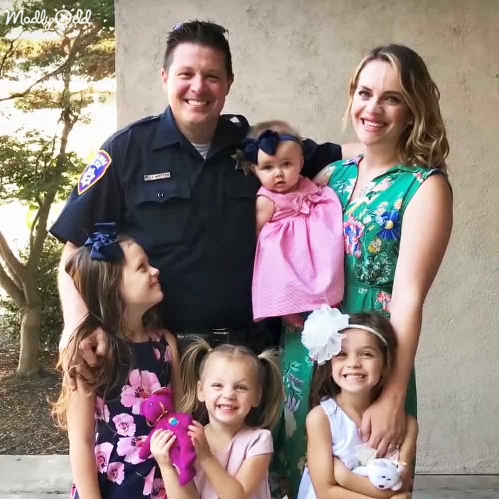 A family portrait with an adult male police officer, two middle-aged adults, a toddler girl, and two young girls outdoors with trees and warm lighting suggesting late afternoon in spring or summer.