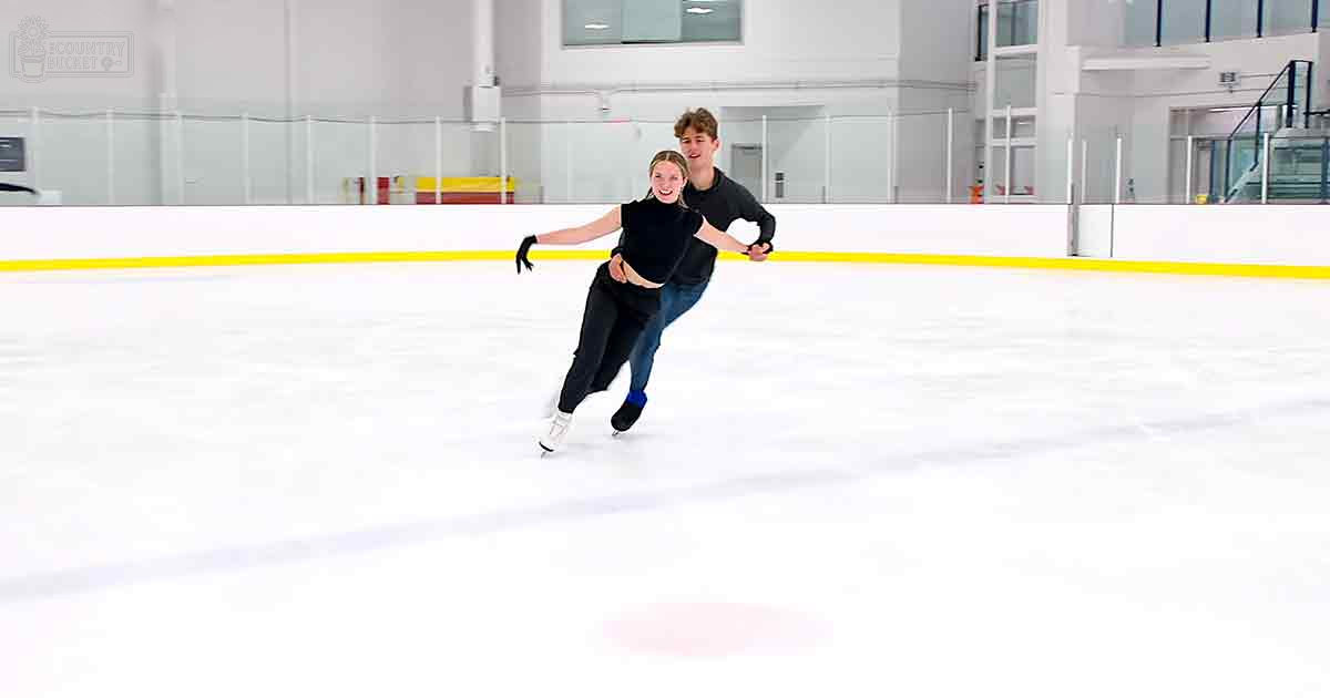 A couple skating together in an indoor ice rink with natural light streaming through windows.
